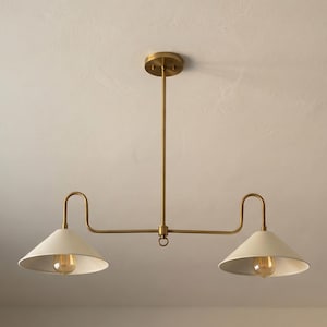 May include: A brass-toned, two-light pendant light fixture. The fixture has a central rod attached to a circular ceiling mount. Two curved arms extend outward, each holding a white, cone-shaped shade with a visible lightbulb.