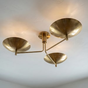 May include: A brass ceiling light fixture with three bowl-shaped shades. The fixture is mounted on a white ceiling. The shades are arranged on arms extending from a central stem.