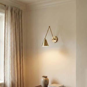 May include: A brass wall-mounted lamp with an adjustable articulated arm and a conical shade. The lamp features a circular wall plate and emits a warm downward light. The fixture is constructed from polished brass with a metallic finish.