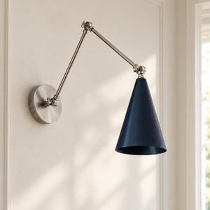 May include: A wall-mounted lamp featuring a brushed nickel finish arm with two adjustable joints and a circular base plate. The light fixture includes a dark navy blue cone-shaped shade that directs light downward from the metal arm.