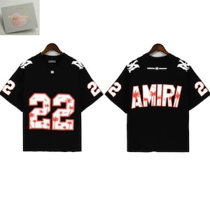 May include: Black short-sleeved shirt with the number "22" in white and red, featuring a palm tree design. The word "AMIRI" is printed on the back in white and red. The shirt is hanging on a wooden hanger.