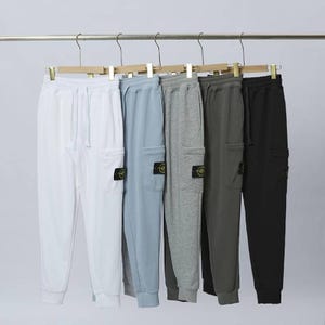 Może przedstawiać: Pięć par spodni dresowych w kolorze białym, jasnoniebieskim, szarym, ciemnoszarym i czarnym, wiszących na wieszaku. Każda para ma kieszeń cargo i naszywkę z logo. Odzież sportowa.