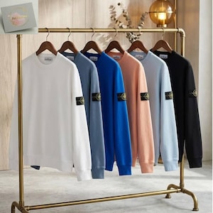 Może przedstawiać: Sześć bluz z okrągłym dekoltem w różnych kolorach, w tym białym, niebieskim i czarnym, wisi na złotym wieszaku. Każda bluza ma naszywkę na lewym rękawie. Bluzy są wystawione na drewnianych wieszakach.