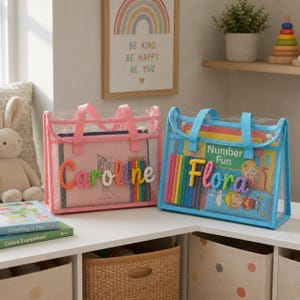 May include: Two rectangular clear plastic tote bags with fabric trim and handles. The pink bag features the name Caroline in colorful yarn letters, while the blue bag displays the name Flora in yarn letters, both filled with books and pencils.