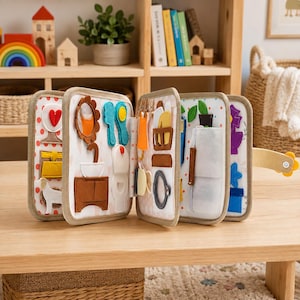 May include: A multi-page felt quiet book featuring various interactive household scenes. The pages display colorful fabric cutouts including a bathroom vanity, a toilet, a mirror, a dog, and various household objects attached with small fasteners.