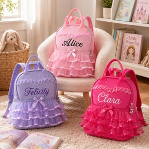 Könnte beinhalten: Drei Rucksäcke in Hellrosa, Lavendel und leuchtendem Pink, jeweils mit gestuften Tüllrüschen, Spitzenbesatz, einer kleinen Schleife und einem in Schreibschrift gestickten Namen: Alice, Felicity und Clara. Jeder Rucksack hat Tragegriffe und Riemen.