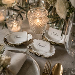 May include: Three natural oyster shells used as place card holders, each containing a rectangular white paper card with deckled edges. The names Olivia, Sophia, and Daniel are printed in black script on the cards, arranged on a table setting.