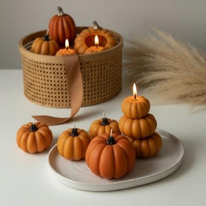 Puede incluir: Velas con forma de calabaza naranja, algunas encendidas, dispuestas sobre un plato blanco y en una cesta de mimbre. Las velas tienen tallos negros y son de varios tamaños. Una cinta y hierba decorativa en el fondo.