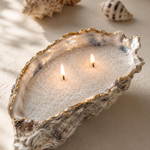 Può includere: Una candela decorativa in una grande conchiglia di ostrica con bordo dorato. La candela ha due stoppini accesi ed è riempita di granuli bianchi. Due conchiglie sono sullo sfondo, creando un tema costiero.