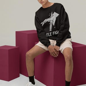 Model wearing a white long-sleeve shirt in a relaxed seated pose, displaying title fight on the front.