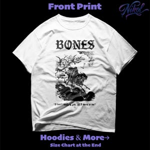 May include: White t-shirt with a black graphic print. The front of the shirt features the word "BONES" above a detailed black and white illustration. Below the graphic is the quote, "I don't need god, all I need is me."