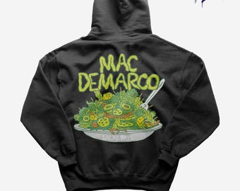 Op de afbeelding: Zwarte hoodie met een afbeelding van een salade, een vork en de gele woorden "MAC DEMARCO" en "SALAD DAYS". De tekst "T-shirts & More" en "Size Chart at the End" zijn ook zichtbaar.
