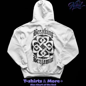 May include: A white hoodie with a black graphic design on the back. The design features the words "Breaking Benjamin" in a gothic font, surrounding a Celtic knot symbol. The hoodie also has a hood and a front pocket.