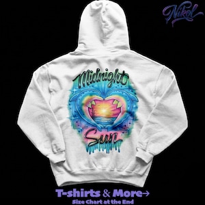 May include: White hoodie featuring a colorful design on the back. The design includes two dolphins forming a heart shape around a sunset scene, with the words "Midnight Sun" in a stylized font. The hoodie also has the text "T-shirts & More" at the bottom.