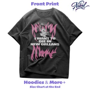 May include: A dark grey t-shirt with a pink and white graphic. The design features the text "I WANT TO DIE IN NEW ORLEANS" and a stylized image. The shirt also displays the brand name "Nikol" in a cursive font.