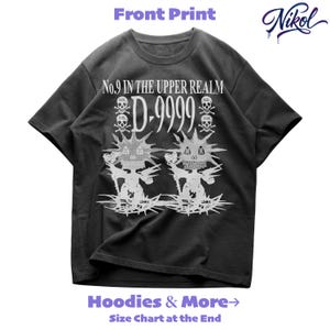 Puede incluir: Camiseta gris oscuro con estampado gráfico blanco. El estampado frontal presenta el texto "No.9 IN THE UPPER REALM D-9999" con calaveras y huesos cruzados y dos figuras de monstruos estilizadas. La marca "Nikol" está en la esquina superior derecha.