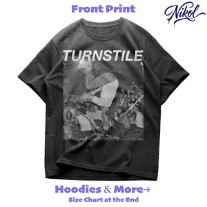 Può includere: T-shirt grigio scuro con la scritta "TURNSTILE" in lettere bianche sopra un'immagine in bianco e nero di una persona a mezz'aria. La maglietta ha anche il testo "Hoodies & More" e "Size Chart at the End". Il logo del marchio "Nikol" è nell'angolo in alto a destra.