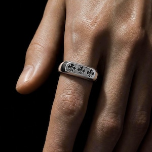May include: A silver-toned band ring featuring a flat, rectangular face engraved with three distinct, small cross-shaped symbols arranged horizontally across the center. The metal surface has a polished finish with dark accents within the engravings.