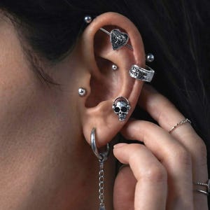 Puede incluir: Un conjunto de pendientes plateados que incluye una barra industrial con un dije de corazón, un aro pequeño con texto grabado, un pendiente de calavera y una cadena colgante. La oreja está adornada con múltiples perforaciones, incluyendo un aro en el lóbulo.