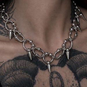 Puede incluir: Un collar de cadena de metal plateado que presenta una serie de eslabones circulares y ovalados interconectados. El collar está adornado con múltiples púas cónicas afiladas unidas a los eslabones circulares, creando un diseño audaz de inspiración industrial.