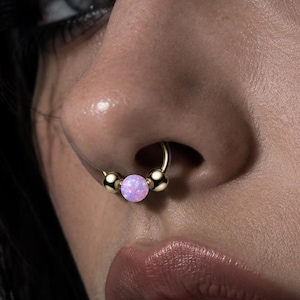Puede incluir: Un anillo de septum circular en tono dorado con dos cuentas doradas lisas que flanquean una cuenta central esférica iridiscente de color rosa claro. La joya está colocada en el tabique nasal, mostrando un acabado metálico pulido y una piedra redonda.