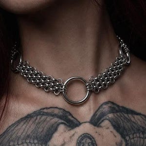 Puede incluir: Un collar tipo gargantilla de cota de malla en tono plateado con un anillo metálico circular central. El collar está construido con pequeños eslabones metálicos entrelazados que forman una banda ancha y texturizada alrededor del cuello.