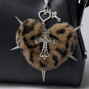 May include: A black leather handbag featuring a heart-shaped leopard print faux fur charm. The charm is adorned with silver-toned metal spikes and a decorative cross pendant, attached to the bag strap with a silver-toned heart-shaped clasp.
