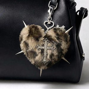 Puede incluir: Un bolso de mano de cuero negro con un dije en forma de corazón hecho de piel sintética con estampado de leopardo marrón y negro. El dije tiene puntas de metal plateado, un cierre en forma de corazón y una cruz plateada con cristales claros.