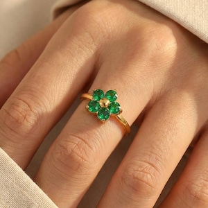 Puede incluir: Un anillo dorado con un diseño floral. La flor está compuesta por cinco piedras preciosas verdes que rodean una esfera central dorada. El anillo se muestra en un dedo.