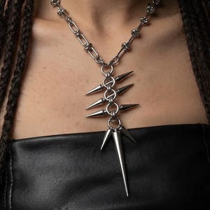 Puede incluir: Un collar de metal plateado con una cadena de eslabones circulares y alargados. El colgante consiste en una disposición vertical de cinco anillos circulares, cada uno adornado con puntas cónicas afiladas que se extienden hacia afuera.