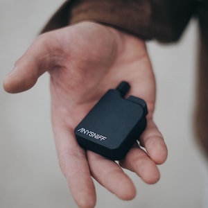 May include: A black rectangular device with the word "ANYSNIFF" printed in white on the side. The device has a small, raised component on top. The device is held in an open hand.