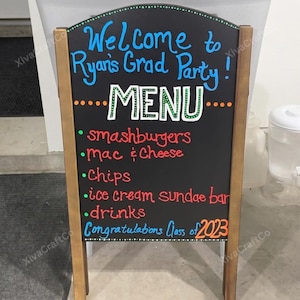 May include: A-frame chalkboard sign with a wooden frame. The black chalkboard features colorful text: "Welcome to Ryan's Grad Party!" and a menu listing smashburgers, mac & cheese, chips, ice cream, and drinks. "Congratulations Class of 2023" is also written.