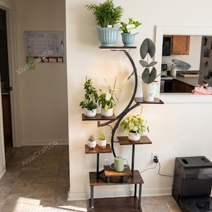 May include: A tall, multi-tiered plant stand with a dark metal frame and wooden shelves. It holds several potted plants of varying sizes, including a large palm, an alocasia, and a small green watering can placed on one of the lower shelves.