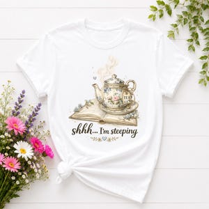 Może przedstawiać: Biały t-shirt z grafiką przedstawiającą kwiatowy czajniczek na otwartej książce, nad którą unosi się para. Poniżej książki znajduje się napis "shhh... I'm steeping" wykonany ciemną czcionką imitującą pismo ręczne, z drobnymi motywami kwiatowymi.