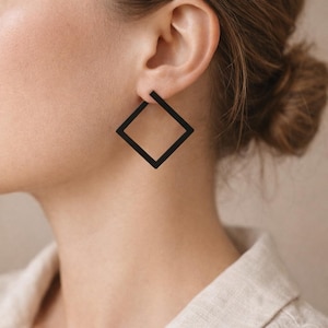 Peut inclure: Gros plan d'une oreille portant une grande boucle d'oreille carrée noire. La boucle d'oreille est de conception géométrique simple, avec un centre creux. La personne porte une chemise de couleur claire.