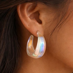 May include: Iridescent hoop earrings. These earrings have a clear, translucent appearance with a subtle rainbow sheen. The earrings are a classic hoop design, adding a touch of modern style. The earrings are worn on the ear.