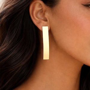 May include: A close-up of a gold-coloured rectangular earring. The earring is a long, slender bar shape. The earring is worn on the ear of a person with dark hair.
