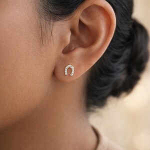 May include: A small gold stud earring shaped like a horseshoe, featuring a series of tiny, clear, sparkling stones set along the curved metal frame. The earring is positioned securely in the earlobe, showcasing a polished metallic finish.