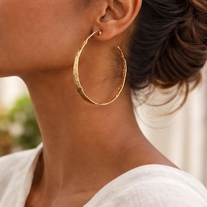 May include: Large gold hoop earrings with a textured, hammered finish. The earrings are a warm gold color and have a simple, elegant design. The hoops are large, making them a statement piece.