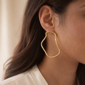 Peut inclure: Boucles d'oreilles dorées de forme abstraite et irrégulière. Elles présentent une surface lisse et polie. L'image est un gros plan mettant en valeur le design fluide et la couleur dorée des boucles d'oreilles.