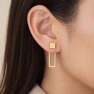 May include: Gold-colored, geometric earrings. Each earring features a small square stud and a rectangular drop. The earrings are shown on an ear, highlighting their modern design and minimalist aesthetic. The earrings are a simple yet elegant accessory.