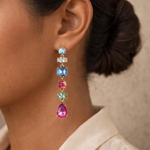 Puede incluir: Un pendiente dorado con una serie de gemas de colores facetadas. El pendiente presenta una piedra azul claro, transparente, azul, rosa, verde y una gran piedra en forma de lágrima magenta. El pendiente se lleva en la oreja de una persona.
