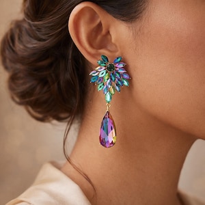 May include: A close-up of a colorful earring. The earring features a cluster of green and blue stones, with a large, teardrop-shaped iridescent crystal pendant. The earring is gold-toned and is worn on the ear.
