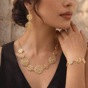 May include: Gold-toned jewelry set featuring a necklace, earrings, and bracelet. The necklace and bracelet have circular, openwork designs. The earrings are a dangling style with two circular elements. The jewelry is worn against a black top.