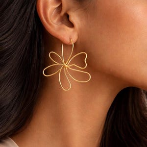 Peut inclure: Une boucle d'oreille créole dorée présentant un motif de fleur en fil métallique avec six pétales arrondis et ouverts. La boucle est formée d'une seule pièce continue de métal qui s'enroule pour créer la forme florale, suspendue à un petit anneau.