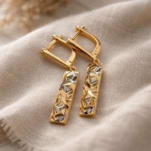 Könnte beinhalten: Ein Paar goldfarbene Ohrringe mit geometrischem Design. Die Ohrringe haben einen rechteckigen Anhänger mit einem strukturierten Muster aus gold- und silberfarbenen Formen. Die Ohrringe werden auf einem beigen Stoff präsentiert.