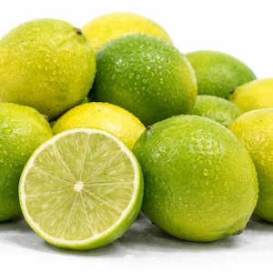 May include: A pile of fresh limes, some green and some yellow, with water droplets on their surfaces. One lime is cut in half, revealing the juicy interior. The limes are arranged on a white surface.