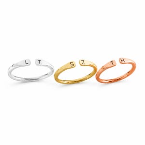 May include: Three open-ended rings in silver, gold, and rose gold. Each ring features a single initial: "L" and "T" on the silver ring, "S" and "Z" on the gold ring, and "T" and "H" on the rose gold ring. The rings are displayed on a white background.