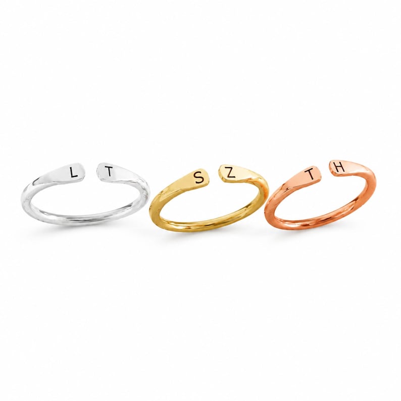 May include: Three open-ended rings in silver, gold, and rose gold. Each ring features a single initial: "L" and "T" on the silver ring, "S" and "Z" on the gold ring, and "T" and "H" on the rose gold ring. The rings are displayed on a white background.