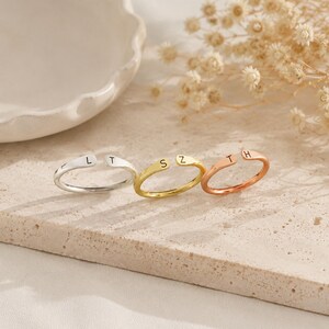 May include: Three open-ended rings in silver, gold, and rose gold. Each ring features a single initial: L, S, and T on the silver ring; S and Z on the gold ring; and T and H on the rose gold ring. The rings are displayed on a neutral-toned surface.